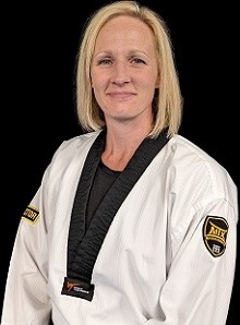 Portrait of a blonde female martial arts instructor wearing a white martial arts uniform, looking confidently at the camera against a black background.