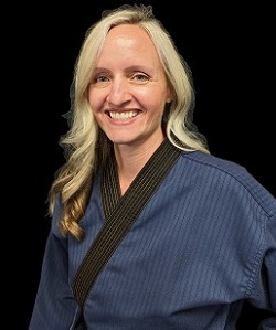 Portrait of a blonde female martial arts instructor wearing a blue martial arts uniform and smiling at the camera against a black background.