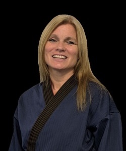 Portrait of a female martial arts instructor with long blonde hair, wearing a blue martial arts uniform, smiling at the camera against a black background.