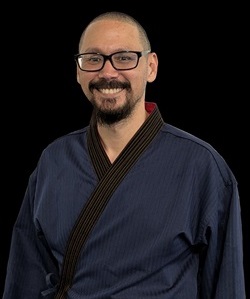 Portrait of a male martial arts instructor with glasses and a goatee, wearing a blue martial arts uniform, smiling at the camera against a black background.