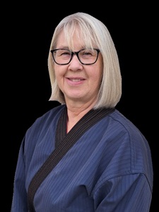 Portrait of a senior female martial arts instructor with blonde hair and glasses, wearing a blue martial arts uniform, smiling at the camera against a black background.