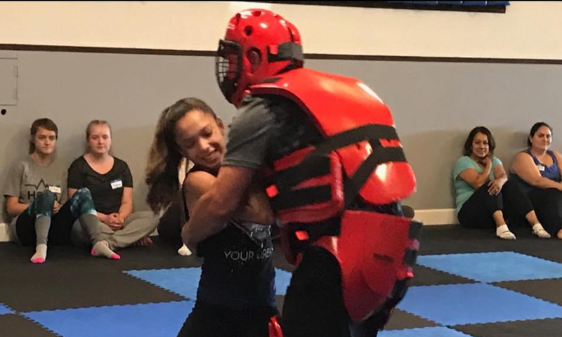 A young taekwondo student practicing self-defense techniques with an instructor wearing red protective padding. Other students sit along the wall watching the training.