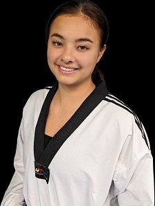 Portrait of a young female martial artist with dark hair, wearing a white martial arts uniform, smiling at the camera against a black background.<br />
