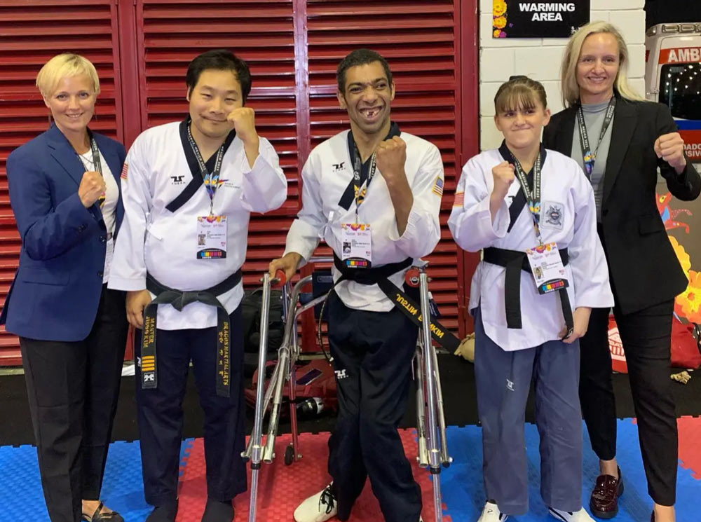 Group photo of adaptive taekwondo athletes and coaches. Two athletes in taekwondo uniforms stand in the center, one using a walker for support. They pose confidently with fists raised, joined by two coaches or officials in business attire. They are standing on a blue and red mat with a "Warming Area" sign and colorful decorations in the background.