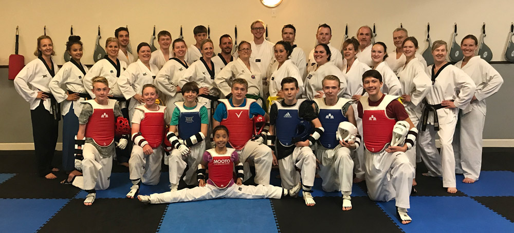 Large group photo of taekwondo team members, including instructors and students of various ages, dressed in white uniforms. Several students in the front row wear sparring gear, including red and blue chest protectors and helmets. A young girl in the center performs a full split in front. The group is gathered on a blue and black mat with training paddles visible on the wall behind them.