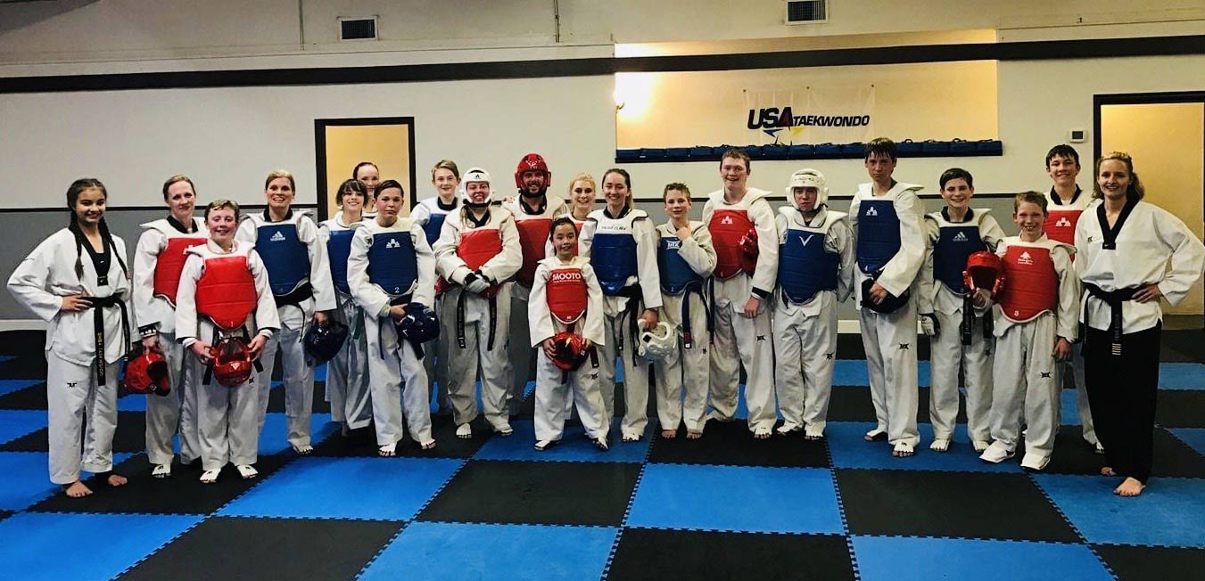 Group of taekwondo athletes and instructors in protective sparring gear, including chest protectors and helmets, posing together on a blue and black checkered mat at a USA Taekwondo training center. The team members vary in age, showcasing a mix of adults and youth. A "USA Taekwondo" sign is visible on the wall in the background.